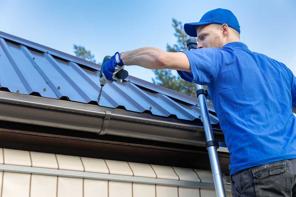A person using gloves to remove leaves and debris from a house gutter on a cloudy day.